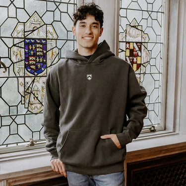 College-aged man wearing the Sigma Tau Gamma embroidered black hoodie, standing in front of stained glass windows with fraternity-style crest designs.