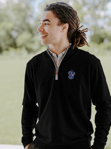 College-aged man smiles and looks off to the side while wearing a black quarter-zip pullover embroidered with a fraternity crest. He stands outdoors in a bright, grassy setting.