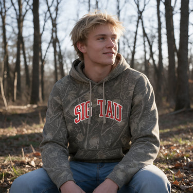 College-aged male wearing the fraternity Deadwood Camo Hoodie with red and white Sigma Tau Gamma lettering, seated outdoors in a wooded setting.