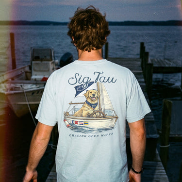 Young man standing on a dock at sunset wearing a light blue t-shirt featuring a golden retriever steering a sailboat with a flag and the phrase “Chasing Open Water” on the back.