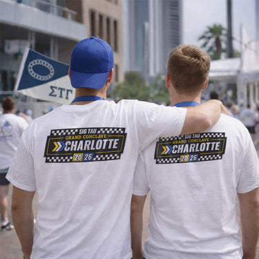 Two young men stand with their backs to the camera at an outdoor rooftop event in front of a city skyline. They are wearing white T-shirts featuring a large back graphic that reads “Sig Tau Grand Conclave Charlotte 2026” with black-and-white checkered flags and yellow and blue racing-inspired accents.