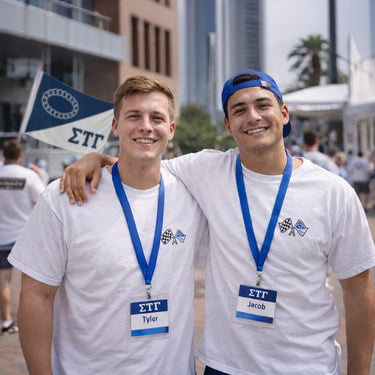 Two young men wearing white event T-shirts and navy lanyards with name badges stand side by side, smiling at the camera. The front of their shirts features a small crossed-flags emblem with checkered and blue flags. Event signage and a city skyline are visible in the background.